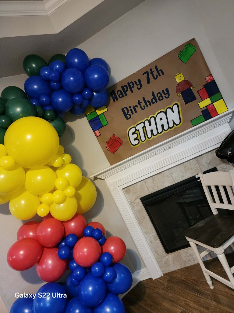 Red, blue, yellow and green balloon garland beside a fireplace with a hand-painted building-brick birthday banner