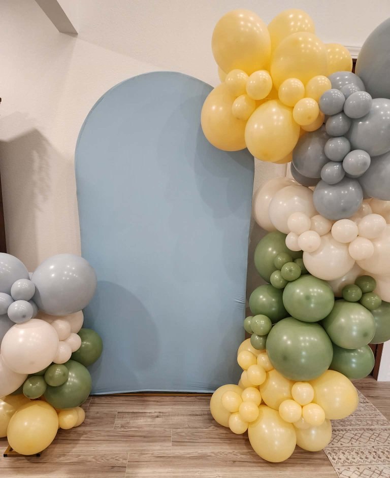 A two-piece balloon set in sage, butter yellow, dusty blue and cream by Belle's Balloons, framing a dusty-blue arch backdrop
