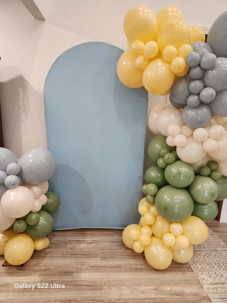 Sage, butter yellow, dusty blue and cream balloon garlands flanking a dusty-blue arch backdrop